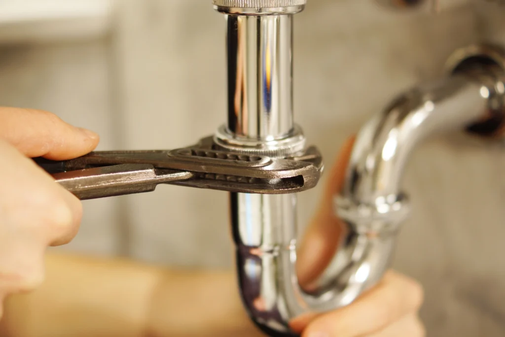 Hands use a wrench to tighten a shiny chrome pipe under a sink, suggesting a plumbing repair. The tone conveys focus and precision.