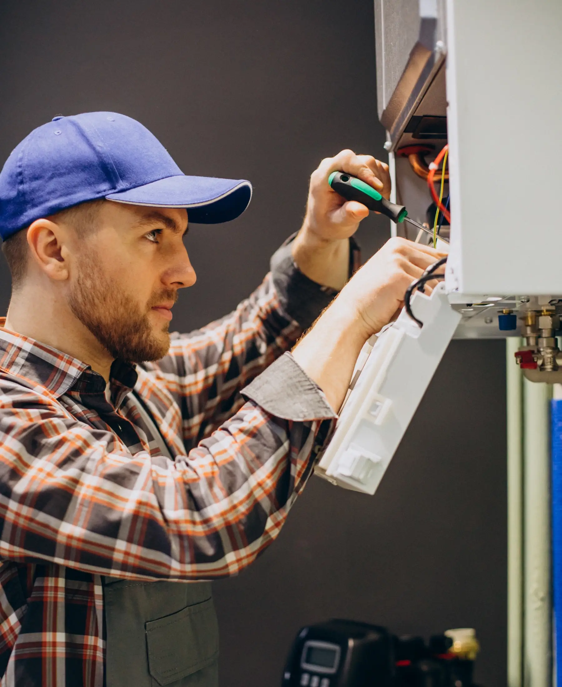 A focused technician in a blue cap and plaid shirt is repairing a boiler. He holds tools and wires, conveying concentration and expertise.